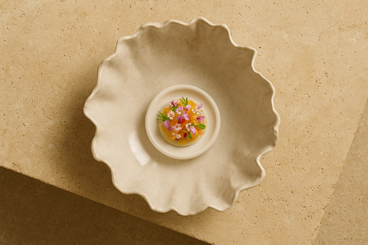 Top view of a gourmet vegetable and edible flower dish served in a sculptural white ceramic bowl, placed on a solid travertine table under soft golden lighting.