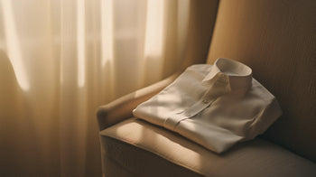 Crisp white Italian dress shirt folded neatly on a textured armchair, softly illuminated by diffused sunlight through sheer curtains—evoking quiet luxury and timeless craftsmanship.