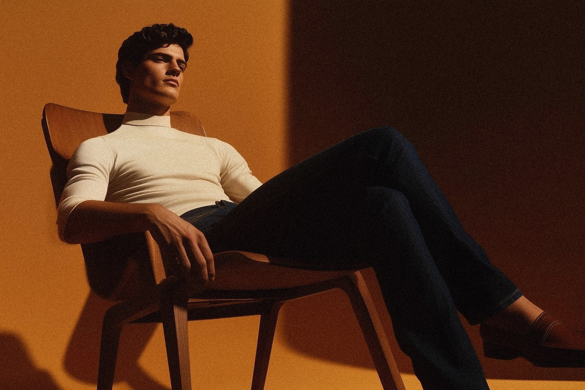 Model wearing custom Maison Alltrueist Japanese denim jeans paired with a camel knit polo, lounging in a walnut chair. Studio-lit with warm shadows for a refined, cinematic mood.