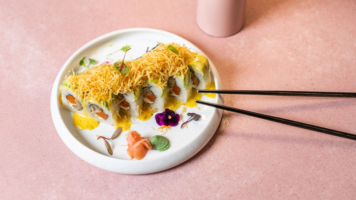Vibrant vegan sushi roll at Mudrá Madrid: avocado-topped maki filled with roasted carrot and king oyster ‘scallop,’ crowned with crispy potato threads and edible blossoms—all plated on pastel pink stoneware.