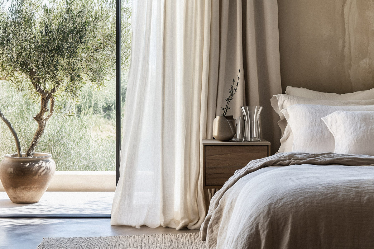 A minimalist bedroom bathed in natural light, featuring an inviting linen-draped bed, soft neutral bedding, and a handcrafted wooden nightstand. A potted olive tree by the window enhances the organic, calming aesthetic.