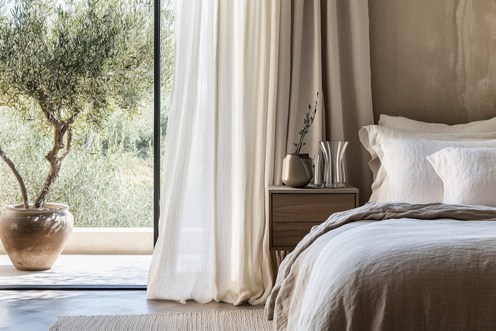 A minimalist bedroom bathed in natural light, featuring an inviting linen-draped bed, soft neutral bedding, and a handcrafted wooden nightstand. A potted olive tree by the window enhances the organic, calming aesthetic.