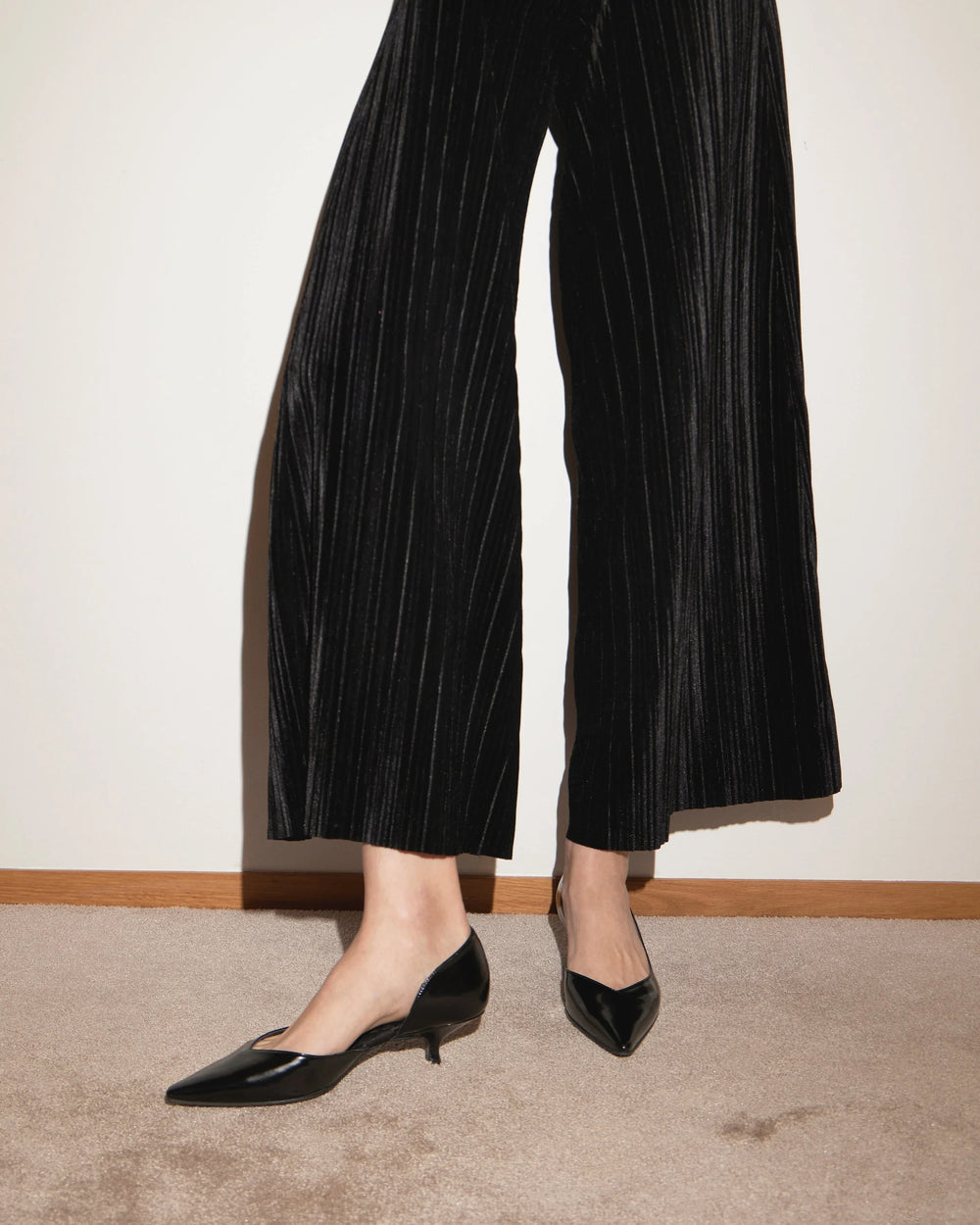 Black pointed-toe kitten heel shoes with wide-leg black velvet pants on beige carpet