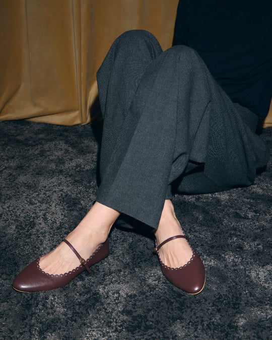 Seated model wearing AERA MINA chestnut Mary Jane flats with grey wide-leg trousers.