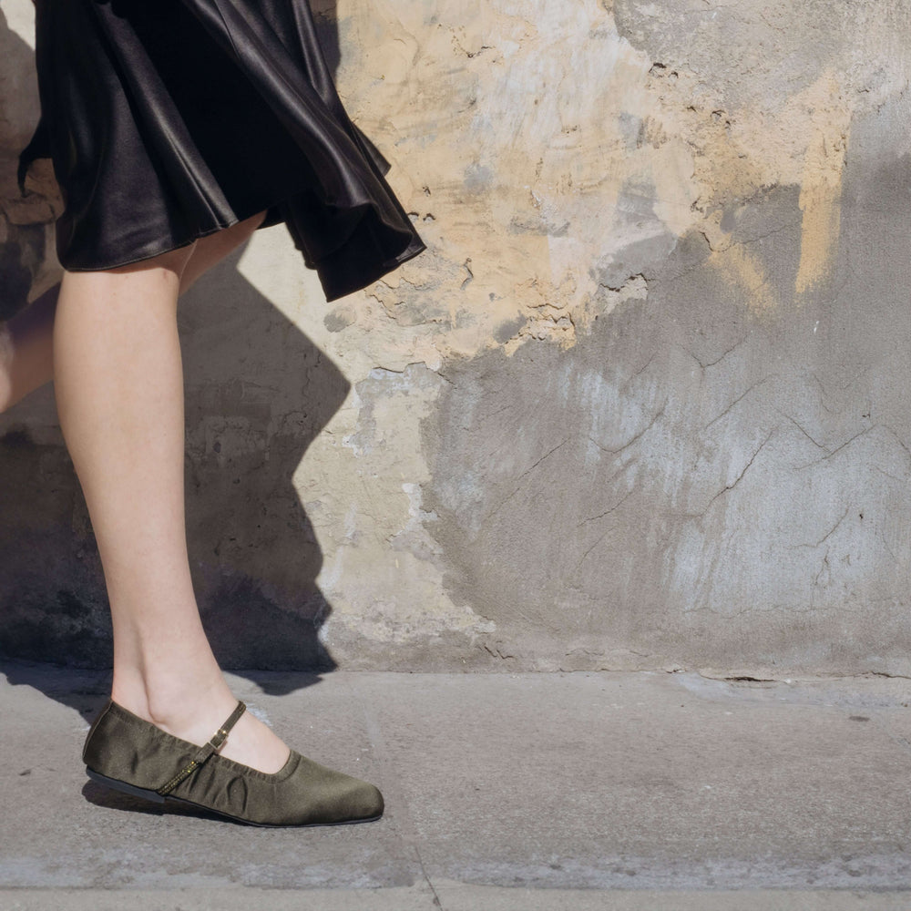 Woman wearing Aurora Military Satin Crystal Mary Jane flats with adjustable strap against a rustic wall background. Vegan, designer, sustainable shoes.