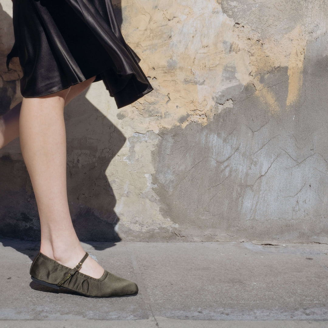 Woman wearing Aurora Military Satin Crystal Mary Jane flats with adjustable strap against a rustic wall background. Vegan, designer, sustainable shoes.