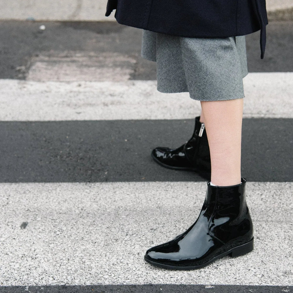 Woman wearing black vegan patent ankle boots crossing the street, showcasing sustainable AERA Charli boots made in Italy.