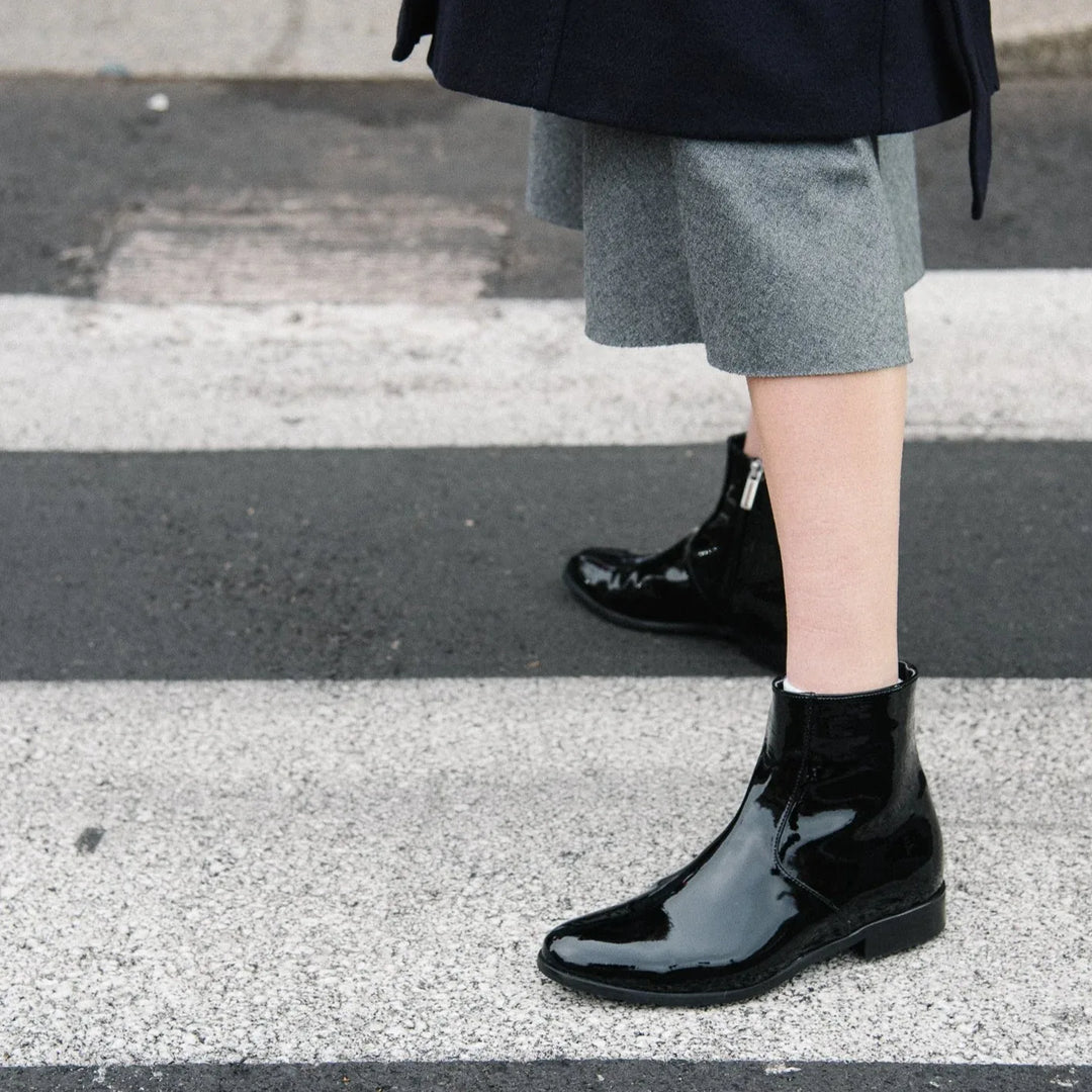 Woman wearing black vegan patent ankle boots crossing the street, showcasing sustainable AERA Charli boots made in Italy.