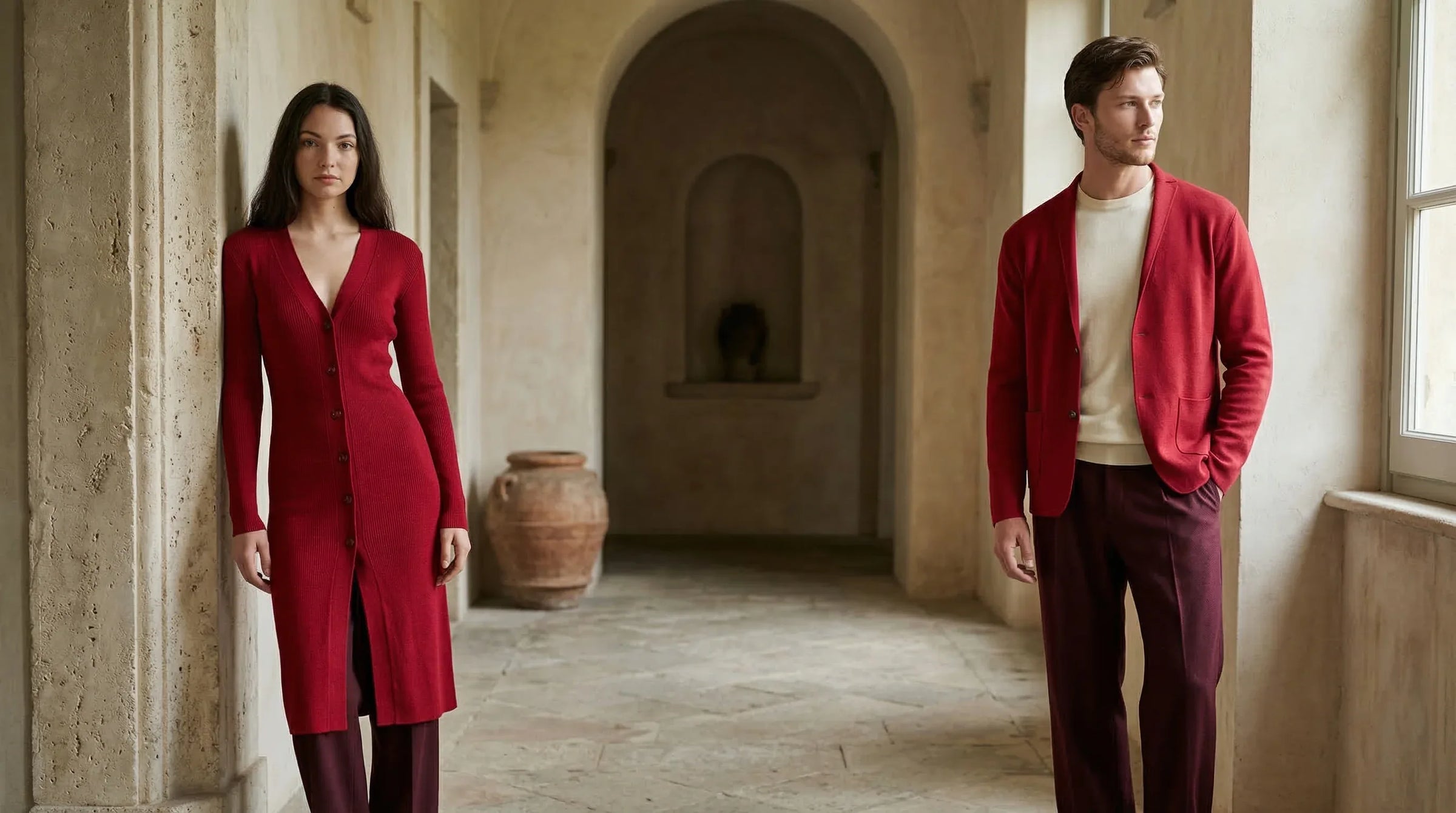 Man and woman in red knitwear and burgundy trousers, standing in a sunlit stone hallway