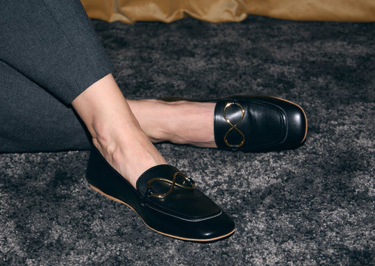 AERA ELLA black loafers with gold-tone infinity hardware, worn with charcoal trousers on a dark carpet.