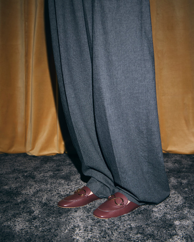Model wearing AERA ELLA chestnut loafers beneath grey wide-leg trousers, in front of gold curtains.