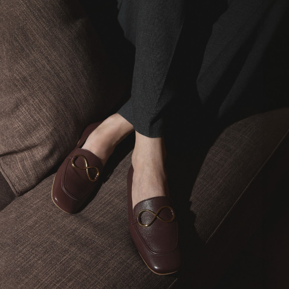 AERA ELLA chestnut loafers with gold-tone infinity hardware, worn by a seated model on a brown sofa.
