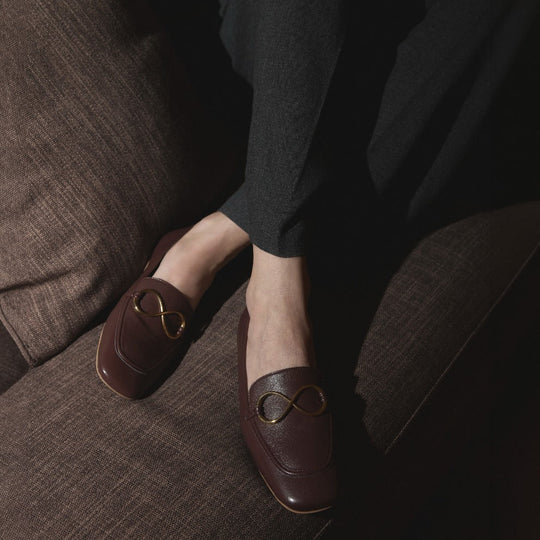 AERA ELLA chestnut loafers with gold-tone infinity hardware, worn by a seated model on a brown sofa.