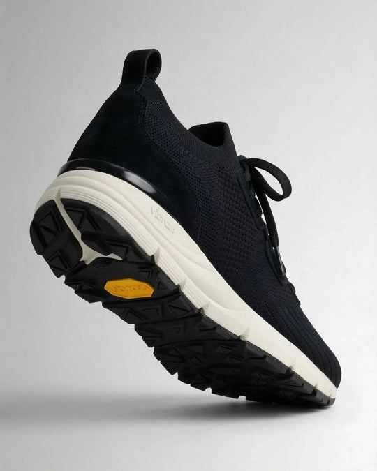 Black suede sneaker with white Vibram sole and textured rubber tread, side angle view