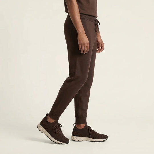Man wearing brown jogger pants with matching knit sneakers on a neutral background