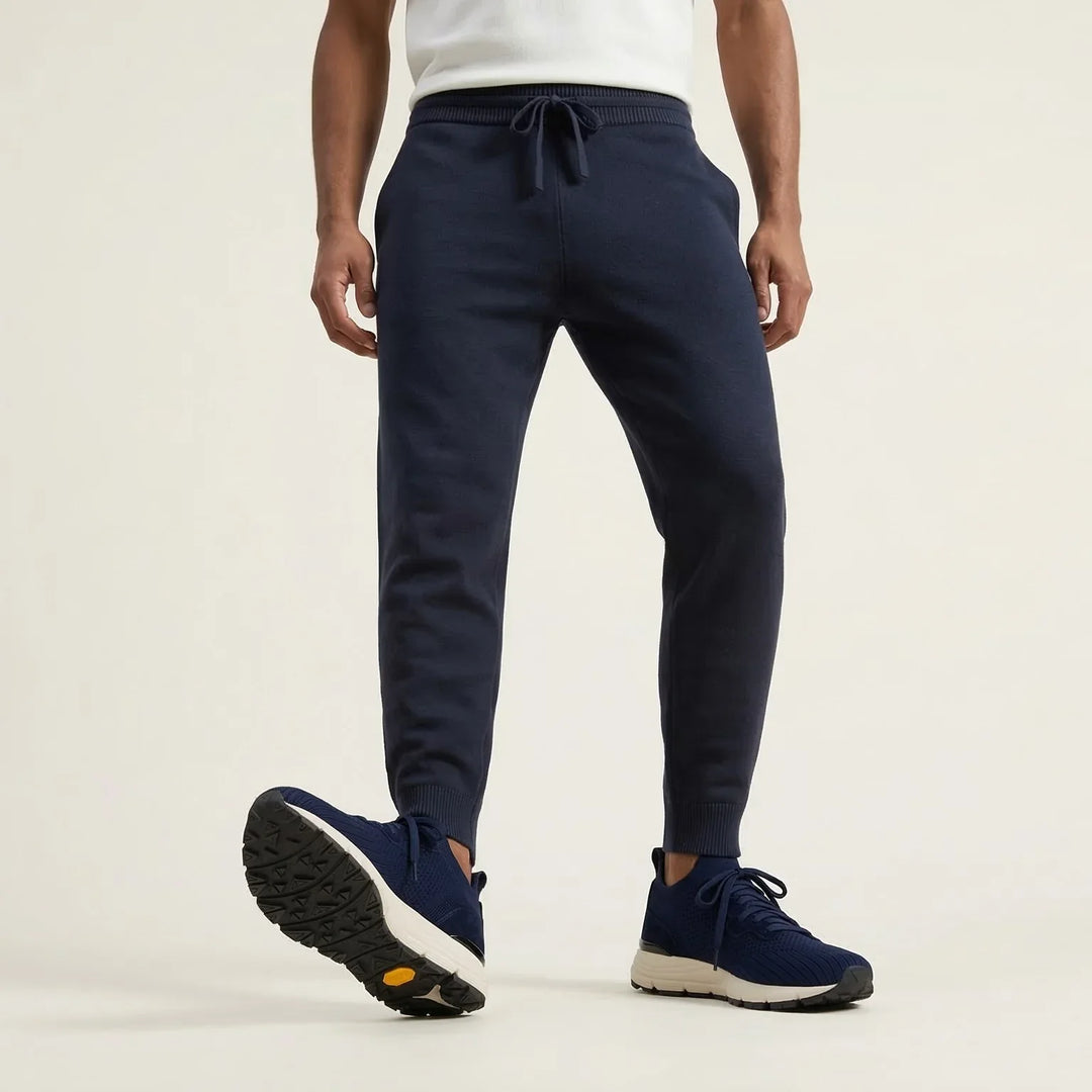 Man wearing navy blue jogger pants and navy sneakers, white t-shirt, neutral background.